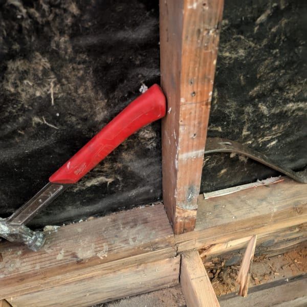 Red-handled hammer wedged against a wooden beam inside an attic or crawlspace.