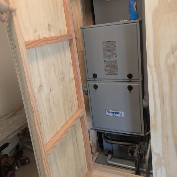 Gray HVAC furnace in a wooden-framed wall opening, with exposed ducting and wiring in a utility space