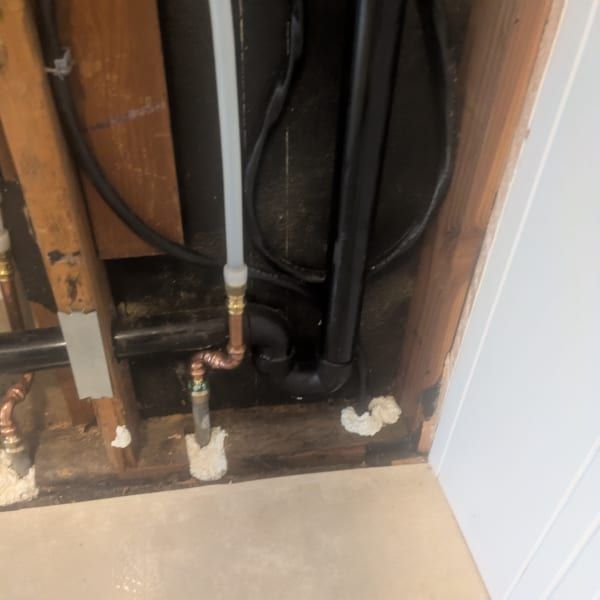 Open wall cavity with exposed plumbing pipes, black tubing, and white foam sealant near a floor edge