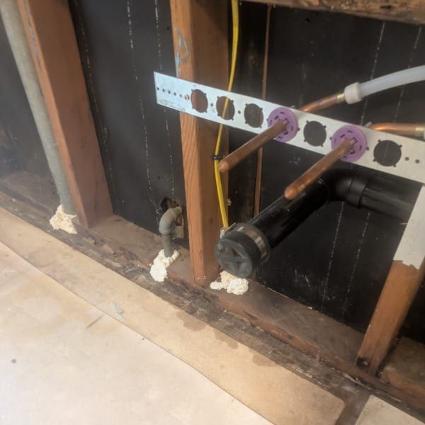 Exposed plumbing and wall cavity under a sink, with pipes, cut drywall, and tiled floor.