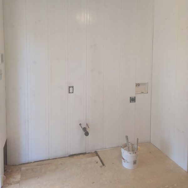 Empty white room with paneled walls, exposed outlets, and a bucket of tools on the floor