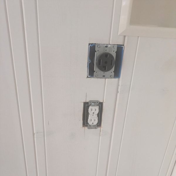Two exposed wall outlets on a white paneled wall, one open electrical box above another standard outlet.