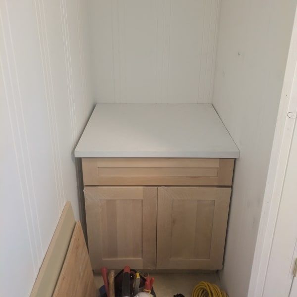 Small wooden cabinet with a gray top in a narrow white alcove, with tools and a board on the floor.