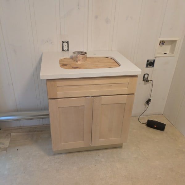 Light wood bathroom vanity with white countertop against a paneled wall, unfinished floor nearby