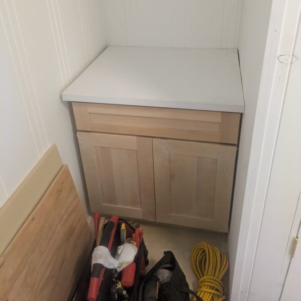 Small cabinet with a white countertop in a narrow utility nook, with tools and an extension cord on the floor.