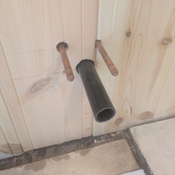 Black pipe and two copper pipes protruding from a wooden wall above a tiled floor.