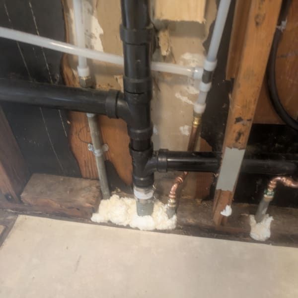 Exposed plumbing in a wall opening, with black and copper pipes and white foam sealing gaps.