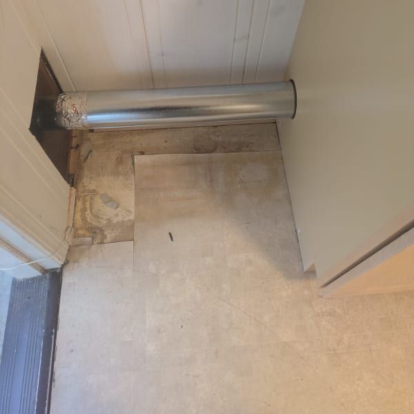 Hallway floor with a silver pipe running along the wall and a damaged patch of flooring near a doorway