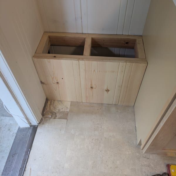 Unfinished wooden cabinet with two sink cutouts in a small room with bare floor and unfinished walls