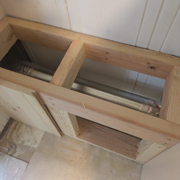 Partially built wooden cabinet frame with open compartments and a metal pipe inside against a white wall