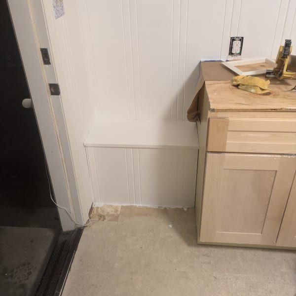 Kitchen corner with white wall paneling, unfinished floor, and a light wood cabinet beside a dark doorway