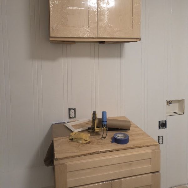 Small unfinished room with beige cabinets and a dresser topped with tools and tape under a wall cabinet.