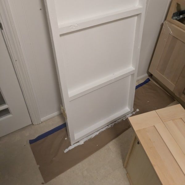White cabinet panel leaning on cardboard in a kitchen, with blue painter’s tape on the floor.