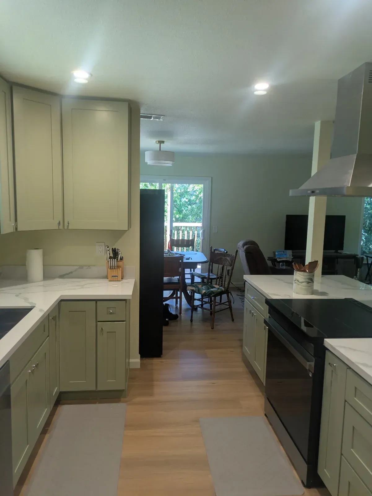Bright kitchen with sage cabinets, white countertops, and a view into a dining room through a doorway.