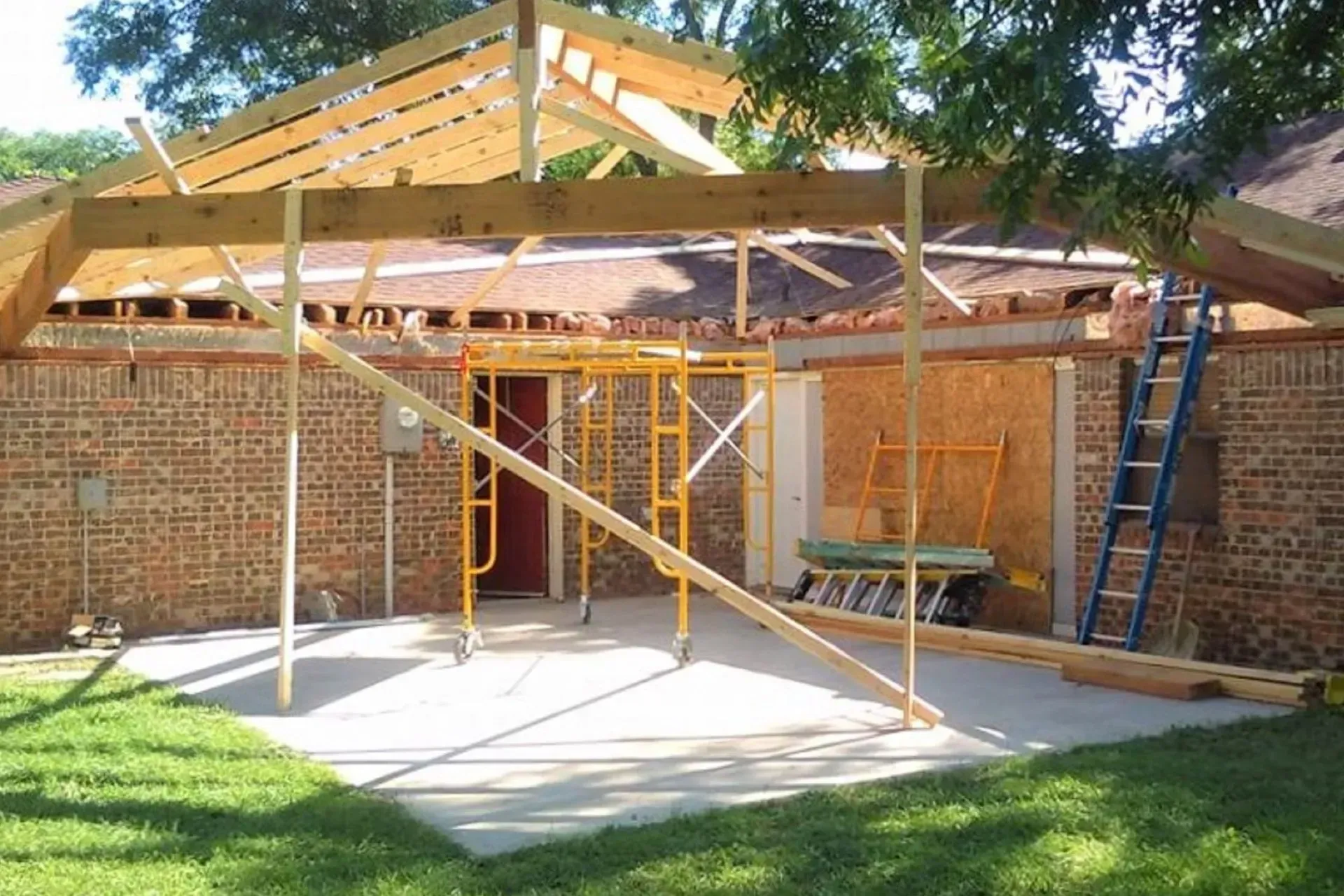 House under construction with brick walls, roof framing, scaffolding, and a ladder in a yard