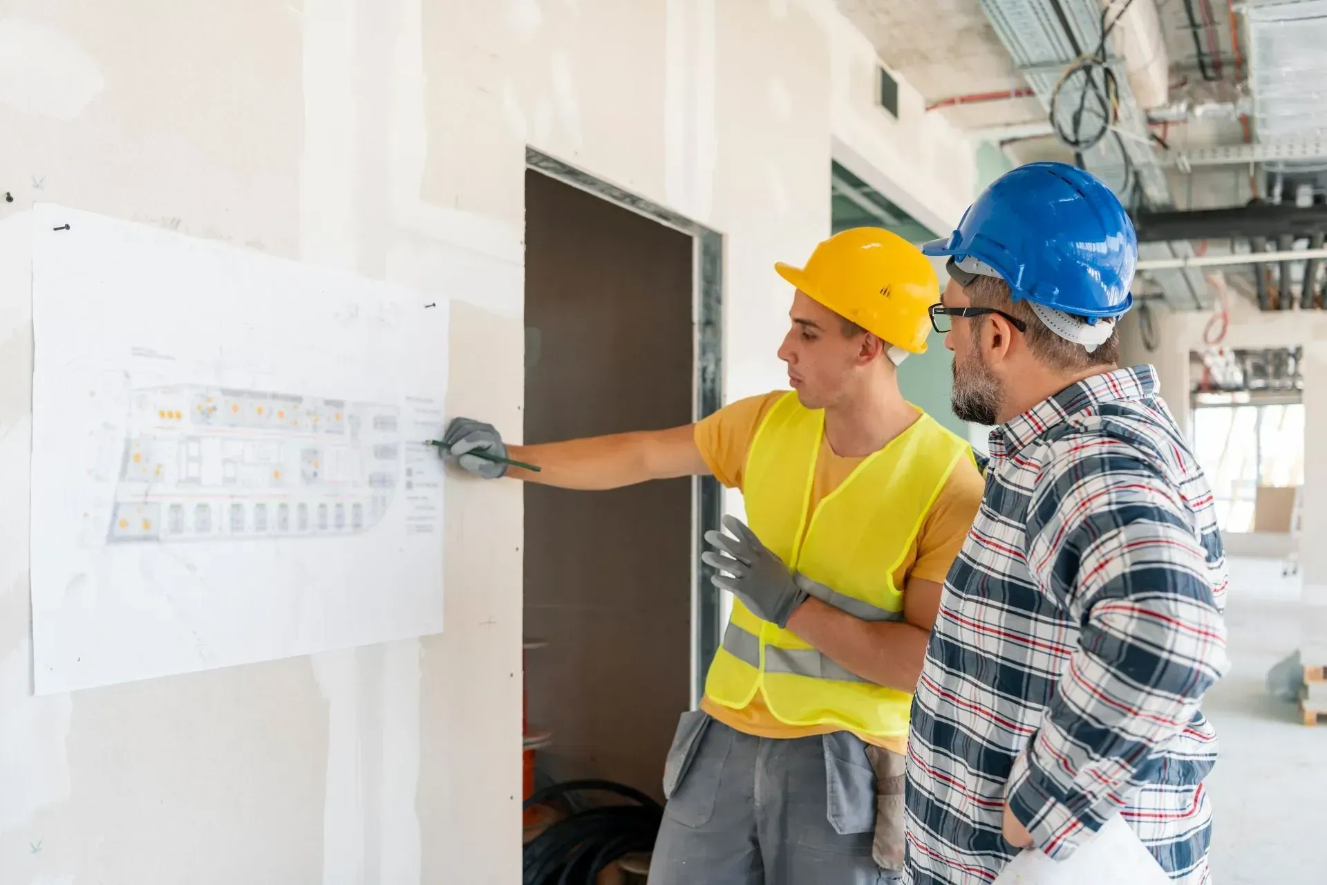 Two construction workers in hard hats review plans on a wall at a job site.