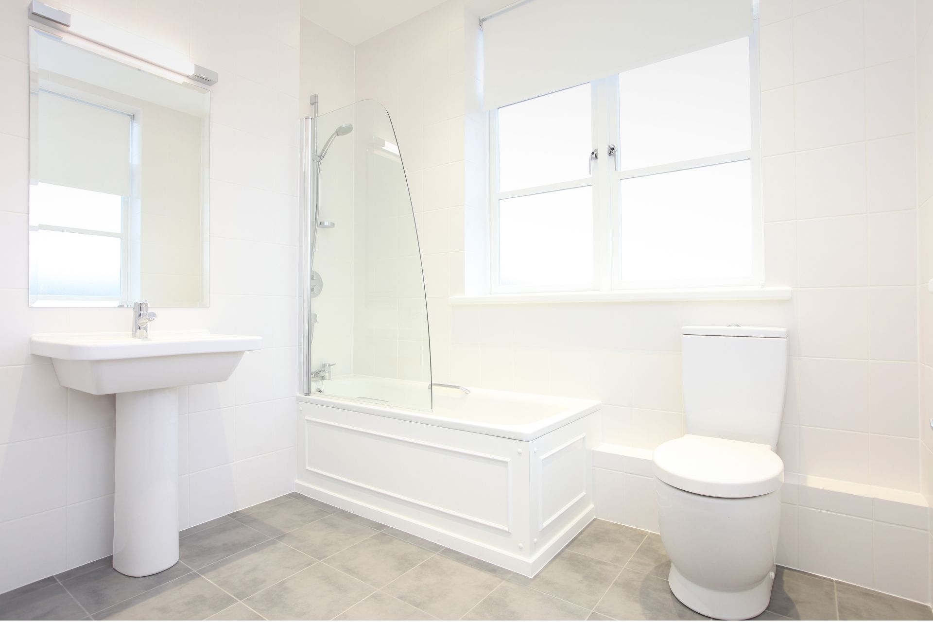 Bright white bathroom with pedestal sink, tub, toilet, large window, and gray tile floor