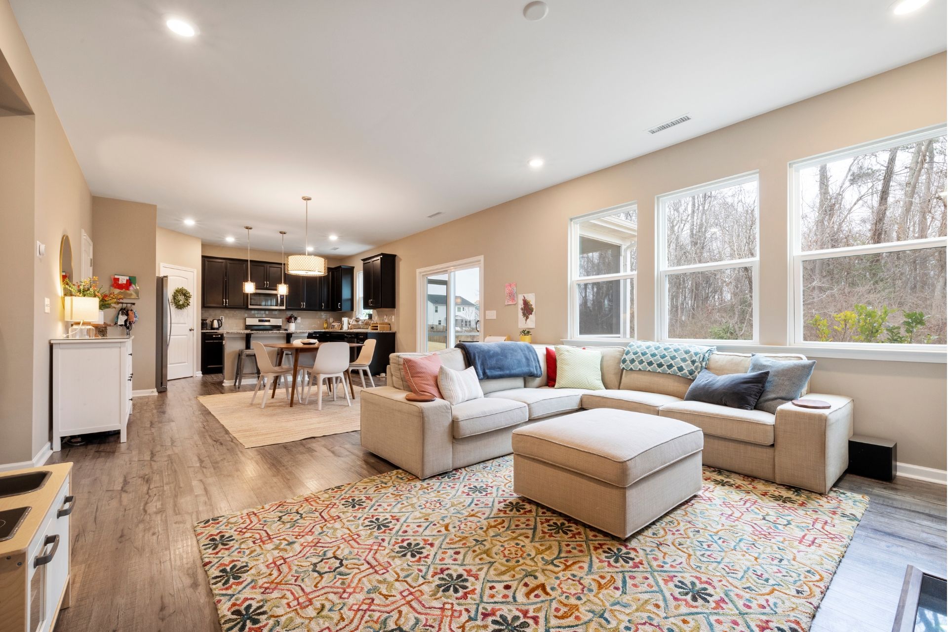 Bright open-concept living room with beige sectional, patterned rug, and kitchen in background