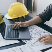 Hands on laptop reviewing architectural plans beside a yellow hard hat on a desk