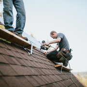 Roofers working on a shingled roof, one kneeling with tools and another standing nearby.