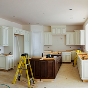 Renovated kitchen with white cabinets, dark island, and a yellow step ladder on unfinished flooring