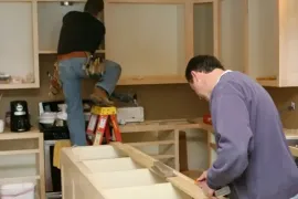 Two people installing cabinets in a kitchen, one standing on a ladder and one working on the countertop.
