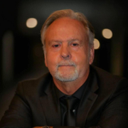 Portrait of a man in a dark suit, facing forward against a blurred dark background with soft lights
