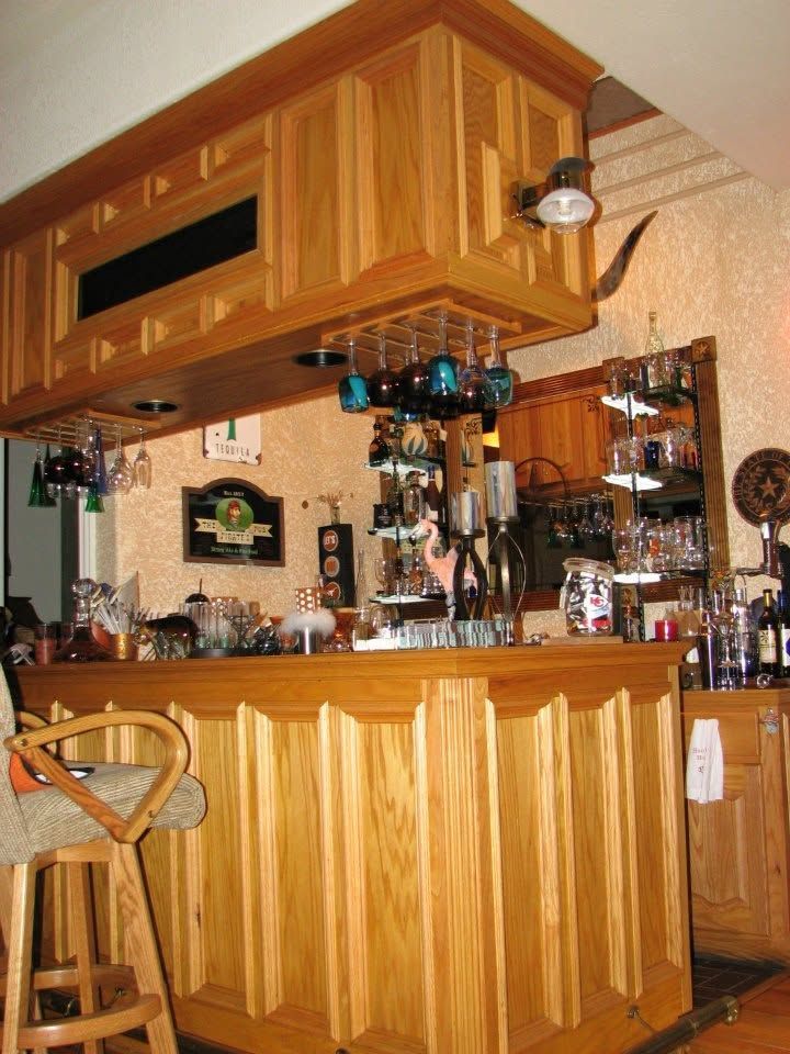 Wood-paneled home bar with hanging glasses, shelves of bottles, and a wooden stool under an overhang