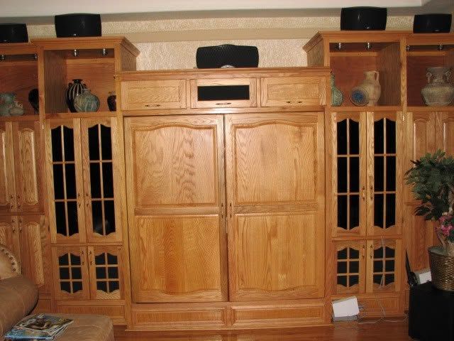 Wooden entertainment center with cabinets and shelves in a living room, flanked by speakers and decor.