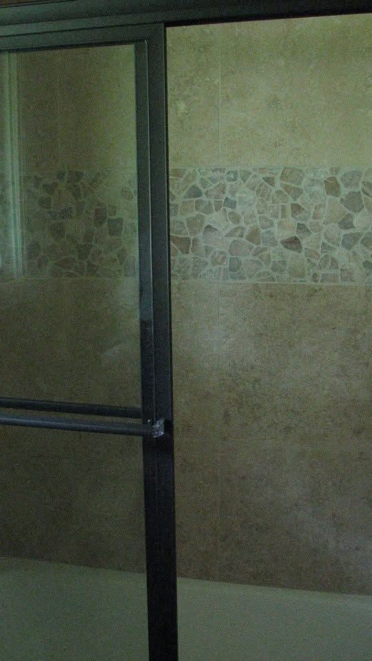 Shower door with black frame beside beige tiled bathroom wall with decorative mosaic band