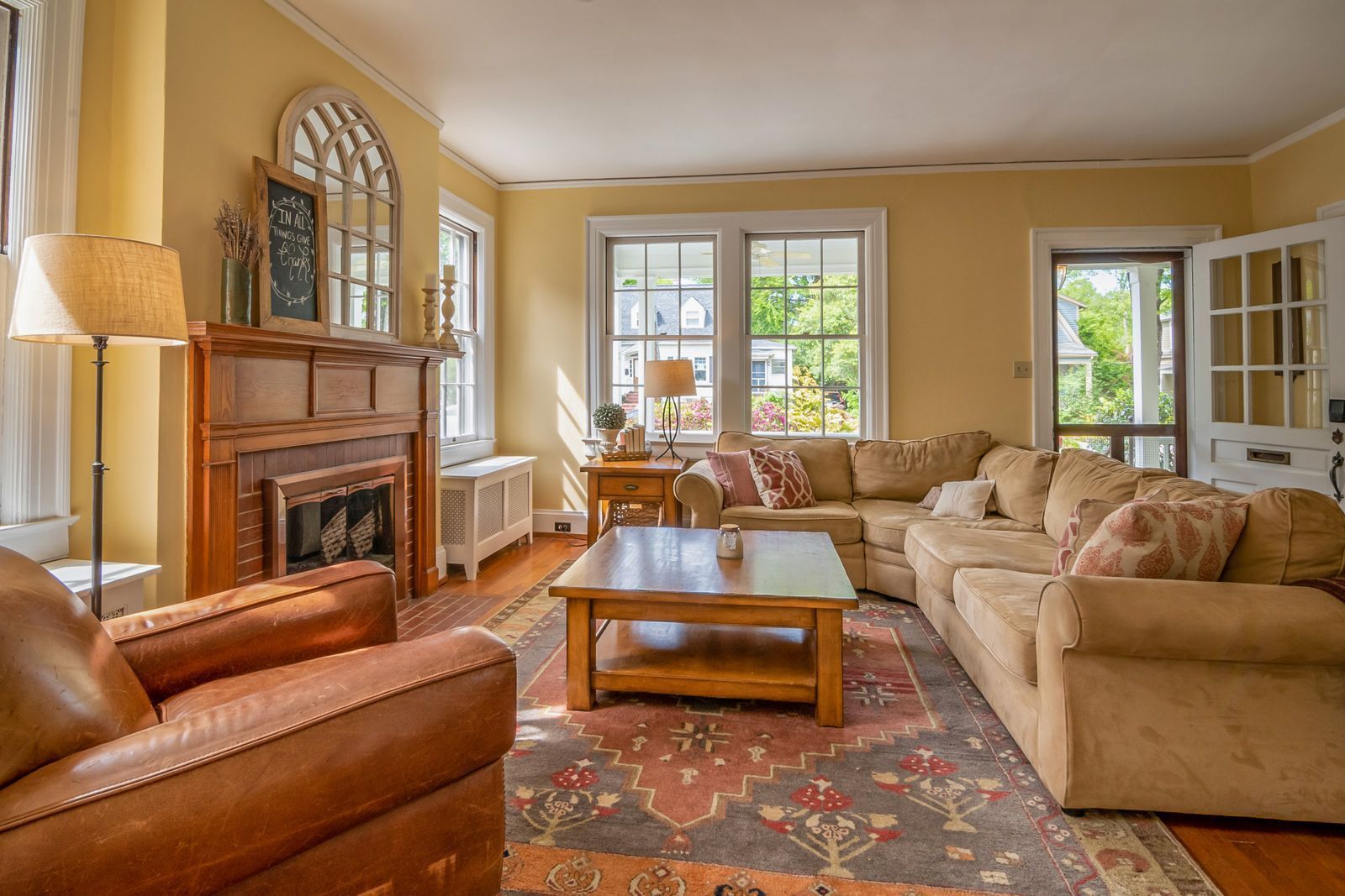 Cozy living room with tan sofas, fireplace, coffee table, patterned rug, and large windows with warm light