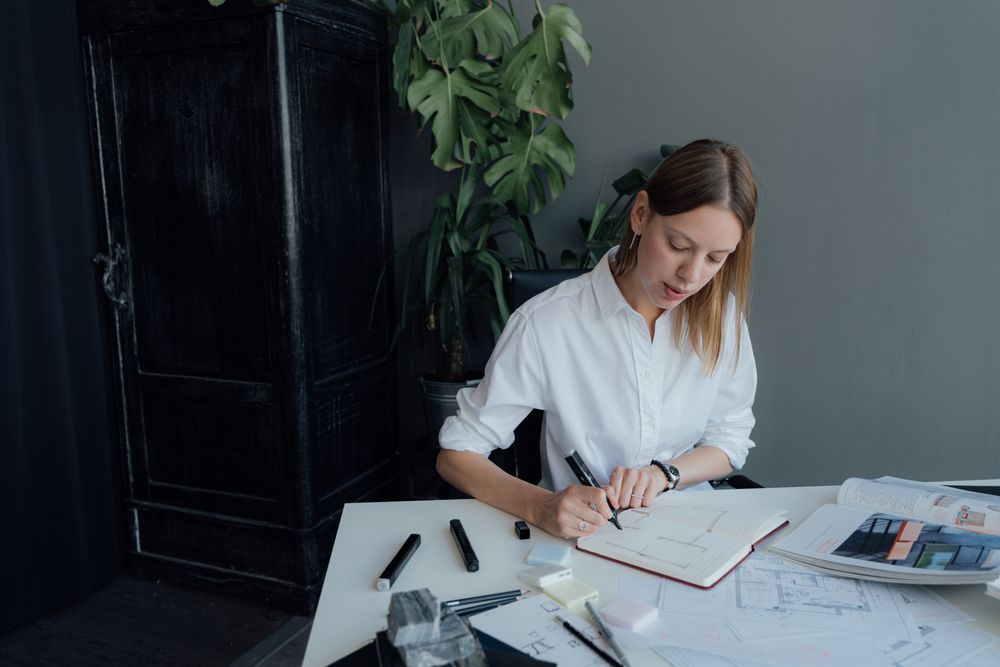 Woman writing in a notebook at a desk with papers, pens, and a laptop in a bright office corner