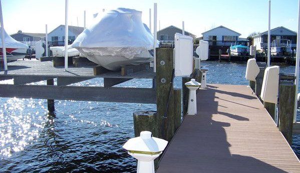 A wooden dock leads to covered boats on lifts, with houses visible across the water in a sunny marina setting.