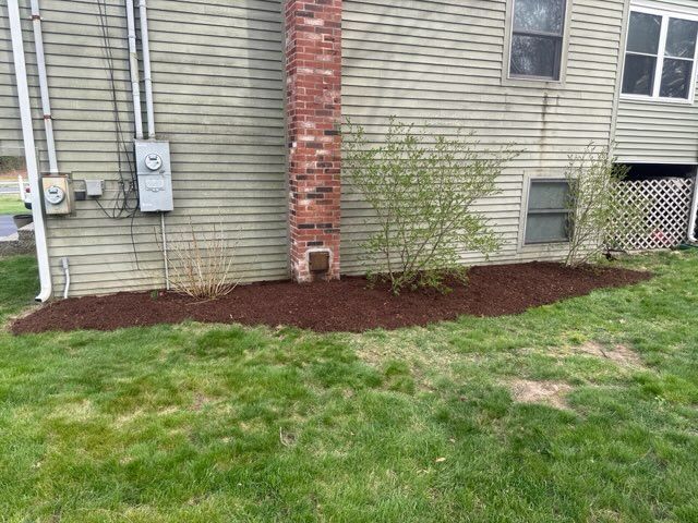 Side of a house with a brick chimney, utility meters, a small shrub, and a mulched bed beside a grassy lawn.