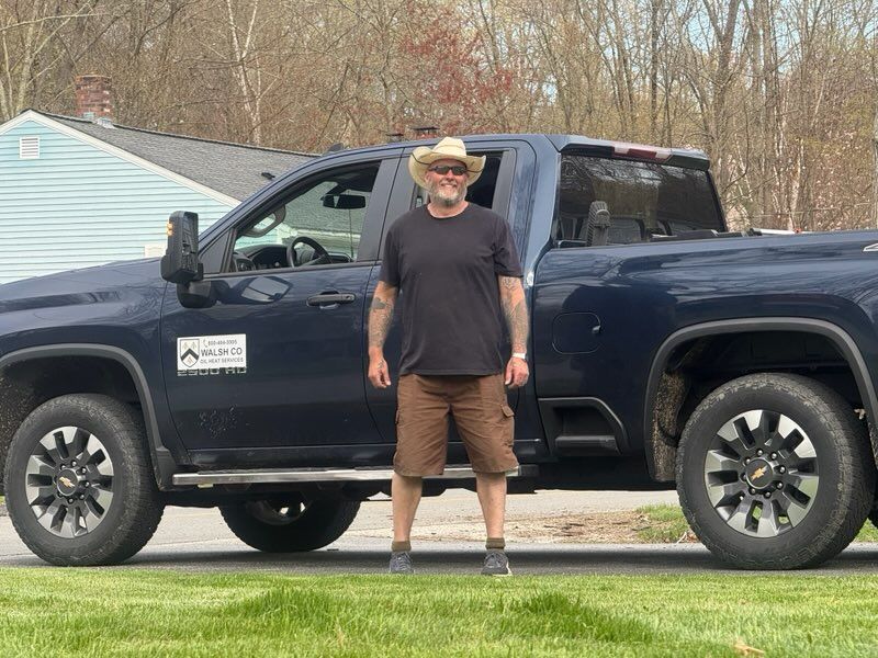 Man standing beside a dark blue pickup truck in a driveway, wearing a hat and black shirt.