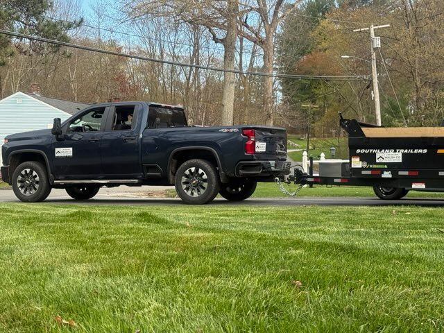 Black pickup truck towing a trailer on a road beside a grassy field, with trees in the background