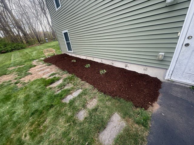 Side yard of a house with fresh mulch, small shrubs, green grass, and a concrete walkway.