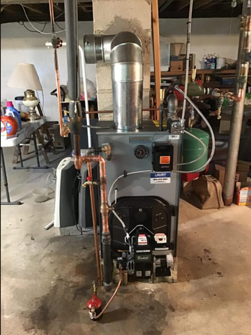 Oil-fired boiler in a basement. Copper pipes and metal chimney vent. Black burner unit and expansion tank.