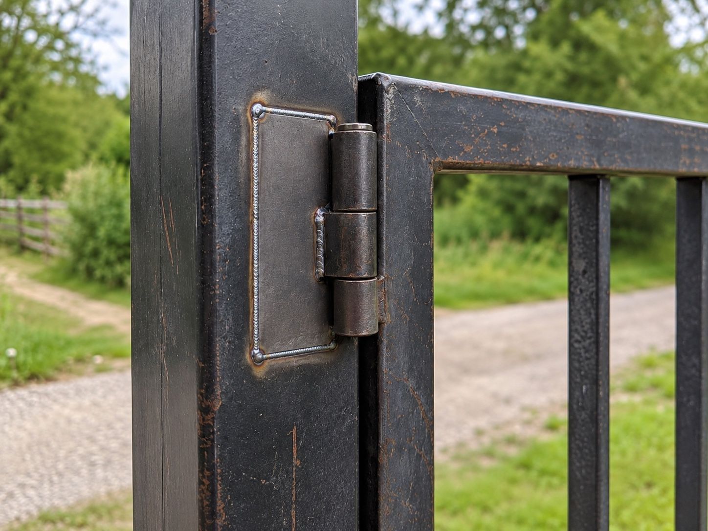 Close-up of a black metal gate hinge on a fence with a gravel path and greenery in the background