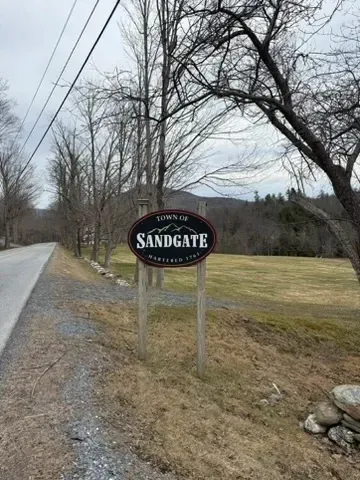 Roadside sign for Sandgate at a rural intersection beside bare trees and a grassy field