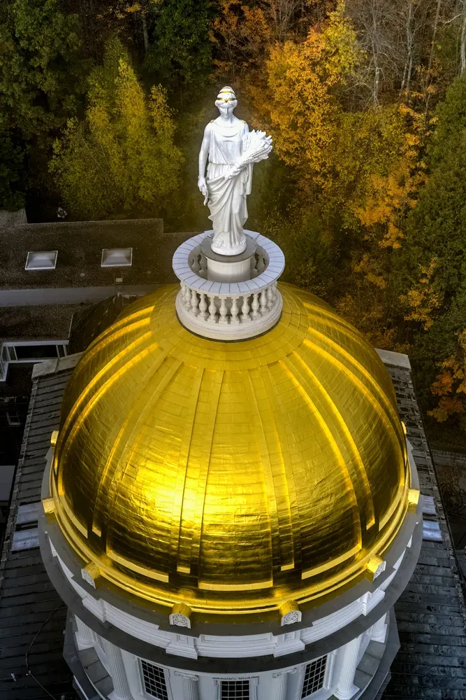 White statue atop a gold-domed building, viewed from above with autumn trees in the background