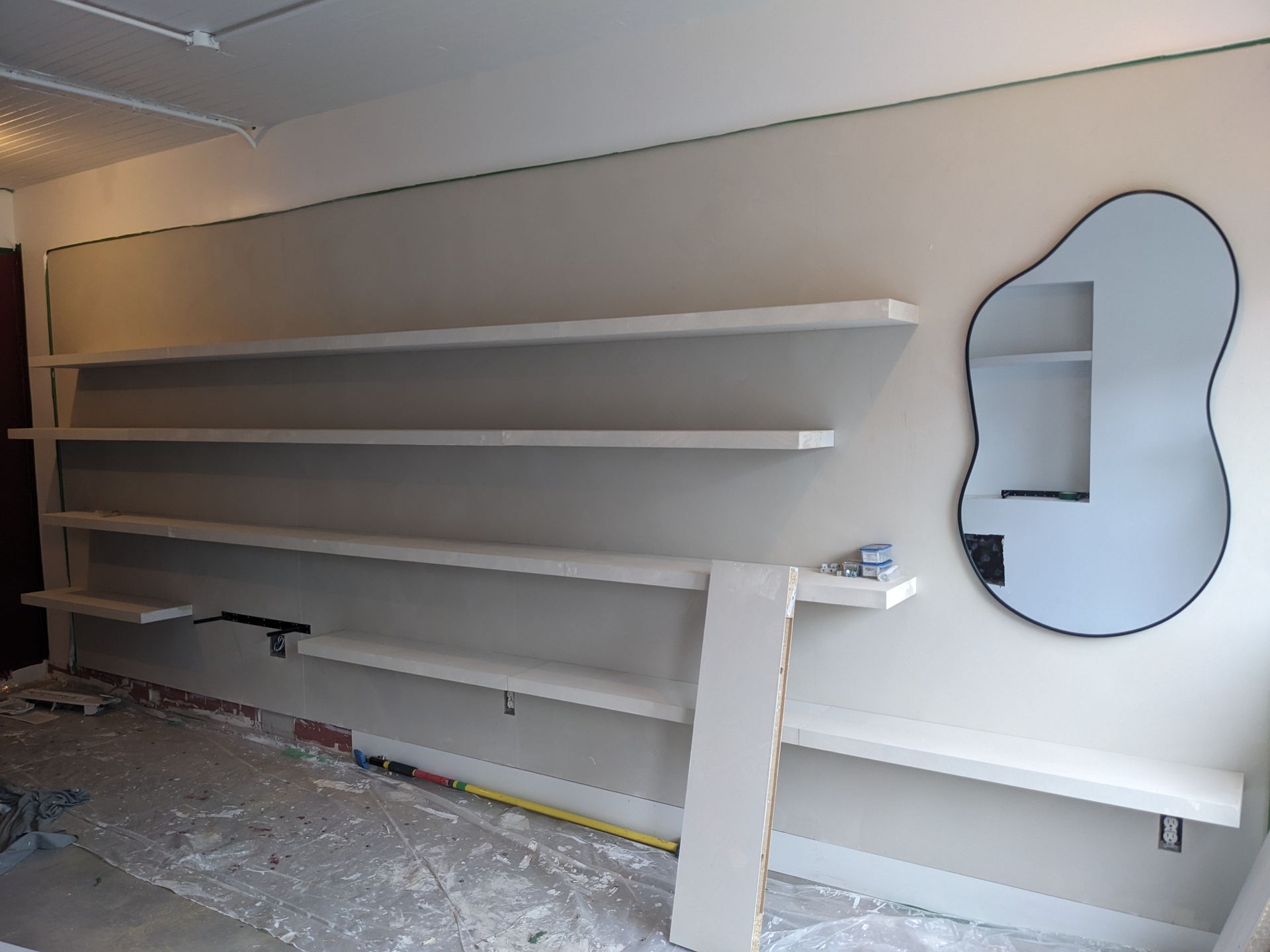 Des étagères sont installées sur un mur avec un miroir abstrait, des matériaux de construction sont posés au sol.
