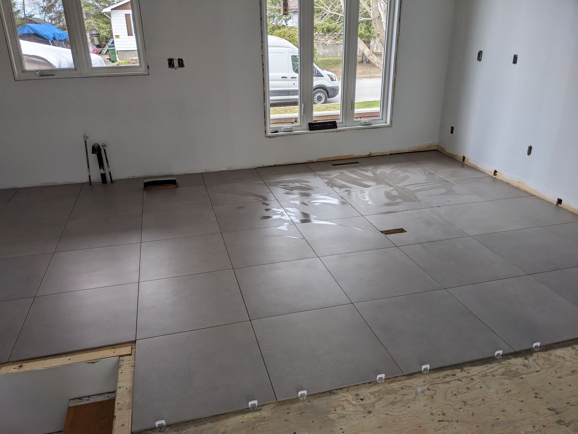 Pièce intérieure avec carrelage gris partiellement posé. Fenêtres et murs blancs visibles.