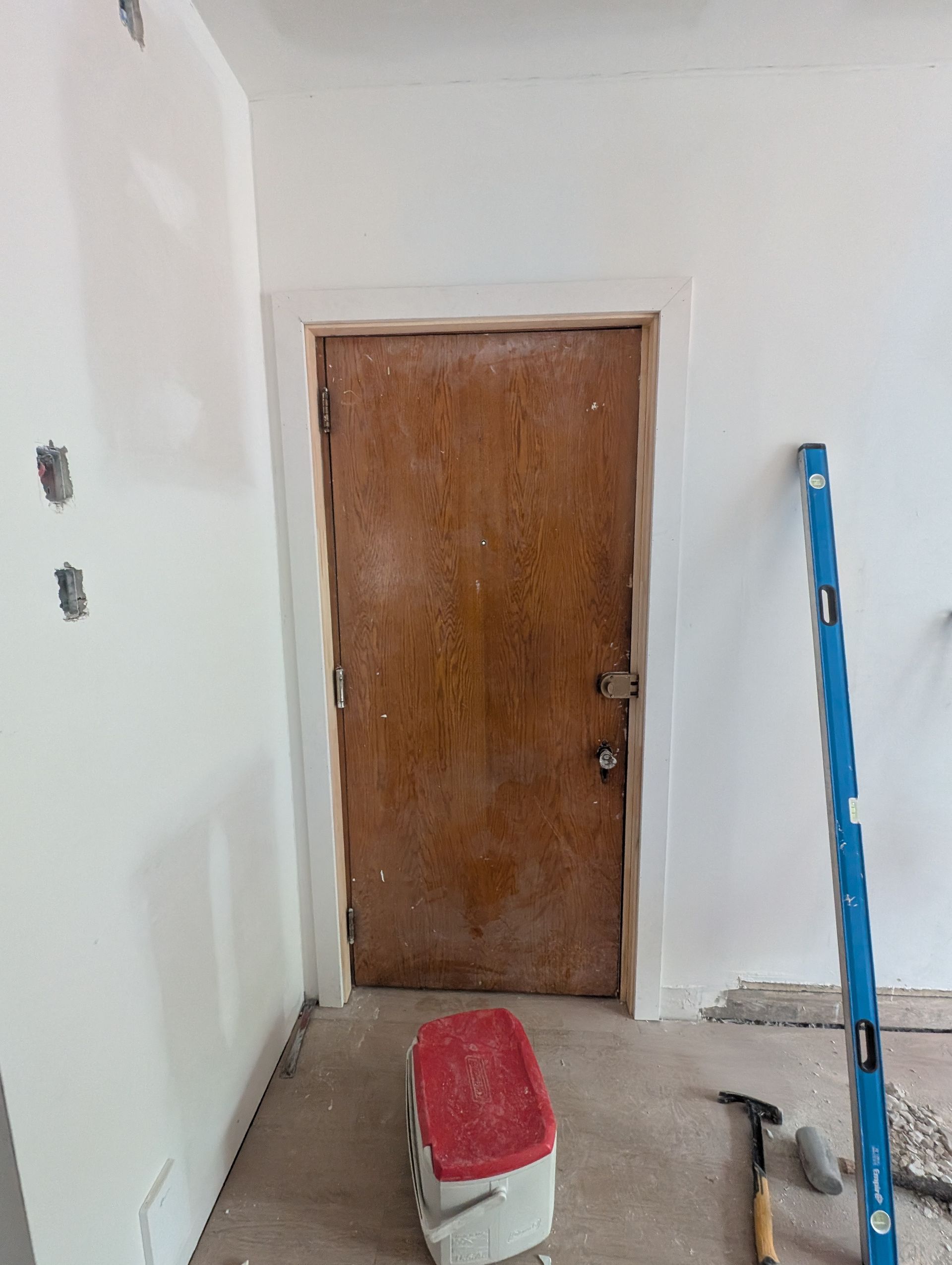 Porte d'entrée avec une porte en bois patinée, des moulures blanches et un niveau bleu appuyé contre le mur.