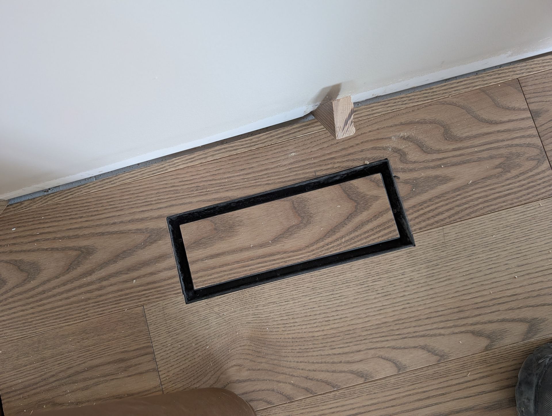 Grille d'aération rectangulaire dans un sol imitation bois, à côté d'un mur blanc orné de moulures et d'un petit morceau de bois.