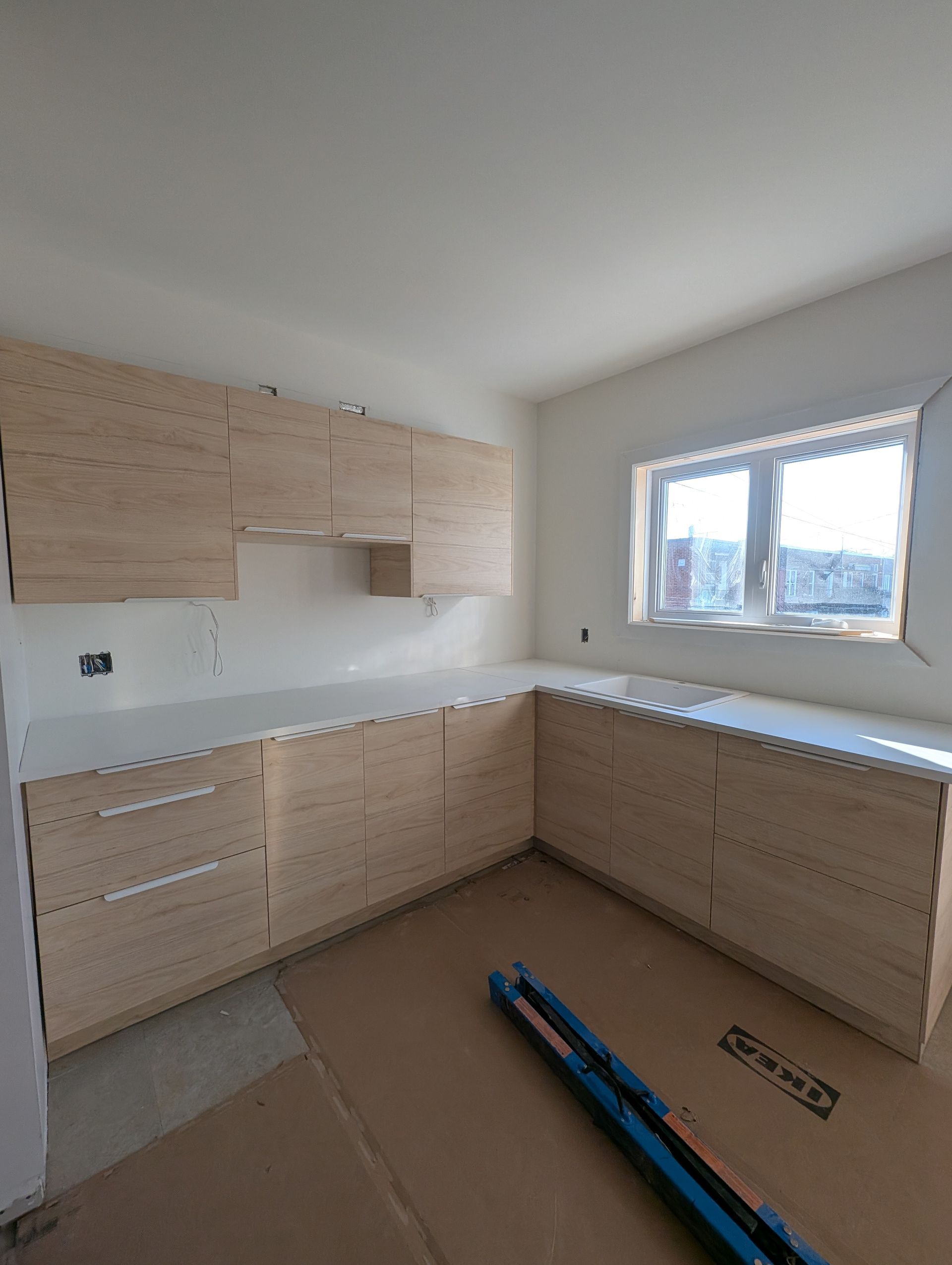 Nouveaux placards de cuisine, finition bois clair, installés dans un coin, avec un plan de travail blanc et une petite fenêtre.