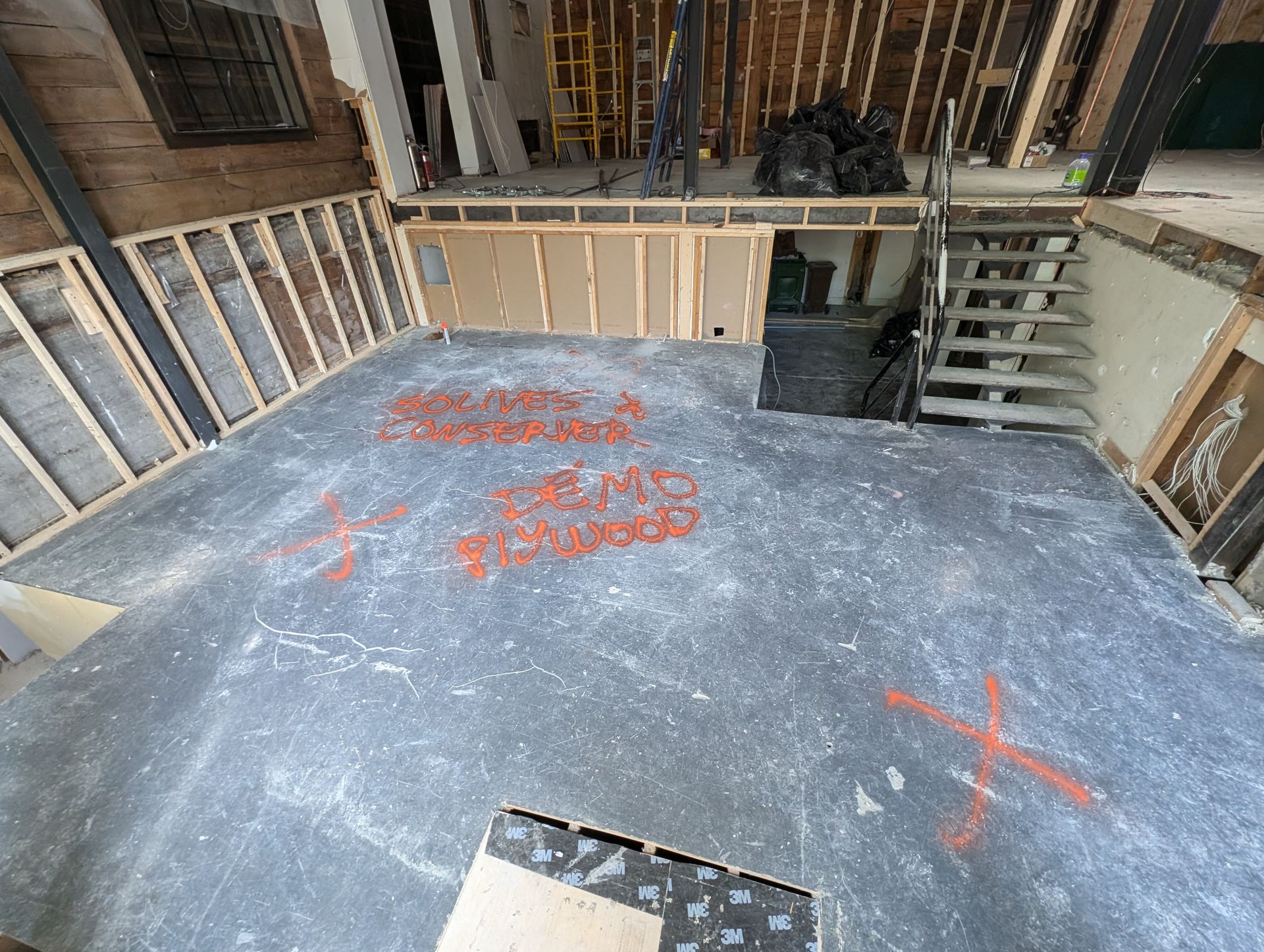 Vue intérieure d'un chantier de construction : sol gris, charpente apparente et escalier. Marquages et inscriptions orange au sol.