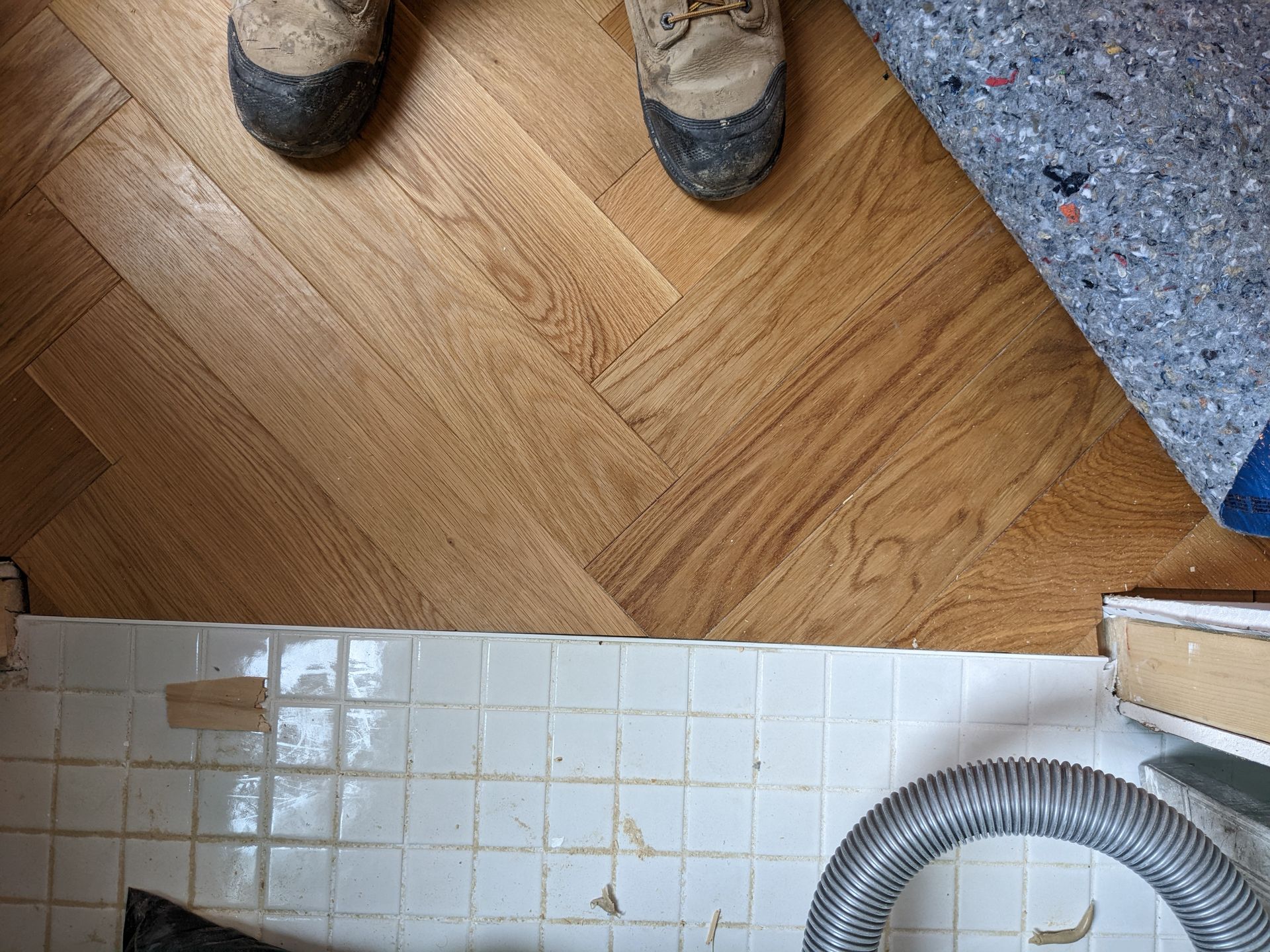 Des bottes de personne sur un parquet, à côté de vieux carrelage et d'une sous-couche de moquette, avec un tuyau d'aspirateur visible.