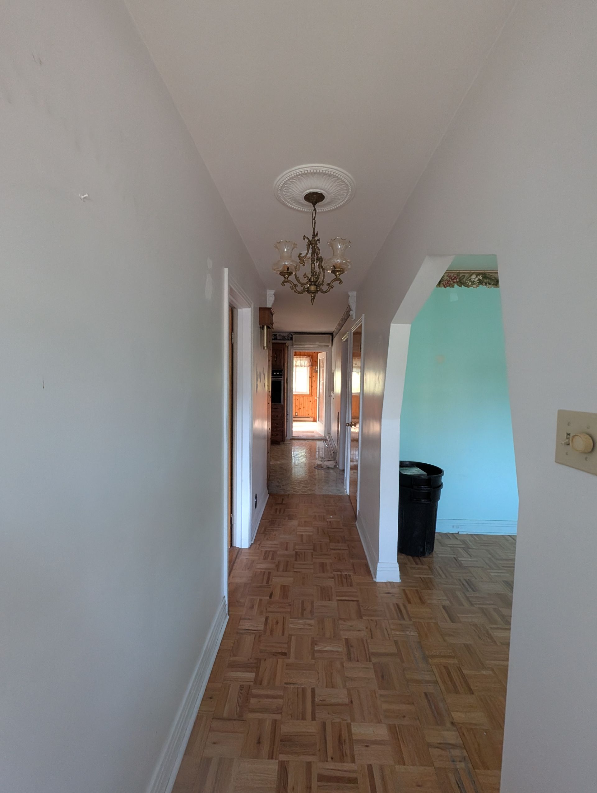 Long couloir avec parquet, murs blancs et lustre. On aperçoit une pièce aux murs turquoise.