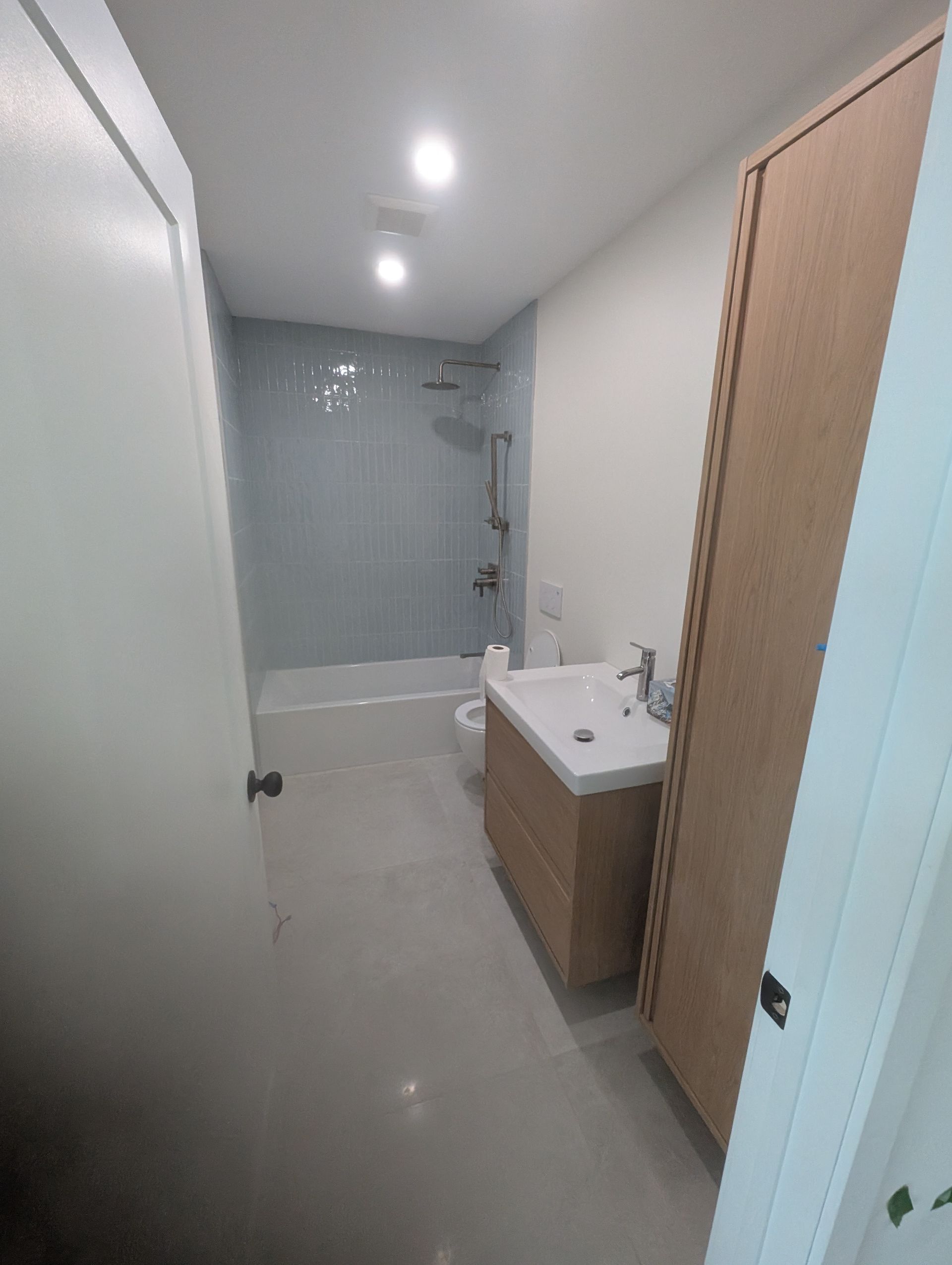 Salle de bains avec murs blancs, meuble-lavabo et armoire en bois clair, et douche carrelée en bleu.